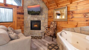 A log cabin interior with a stone fireplace, a television mounted above it, a large mirror, a bathtub, and a patterned rug. - Location Hot Tub Privacy Fireplace Common Pool (Gatlinburg)