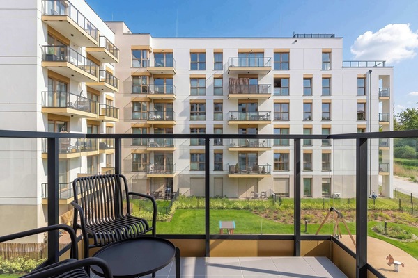 A private balcony with black metal mesh chairs and a matching small table, overlooking a modern residential courtyard. The glass balustrade provides an unobstructed view of the manicured green lawns and pathways below.