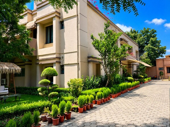 A building with a paved walkway lined with potted plants, a thatched-roof structure, and a clear blue sky. - Vrindavan Staycation (Mathura)