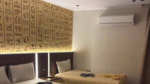 Une chambre d’hôtel avec deux lits, un mur orné d’hiéroglyphes, un luminaire au plafond et un appareil de climatisation.