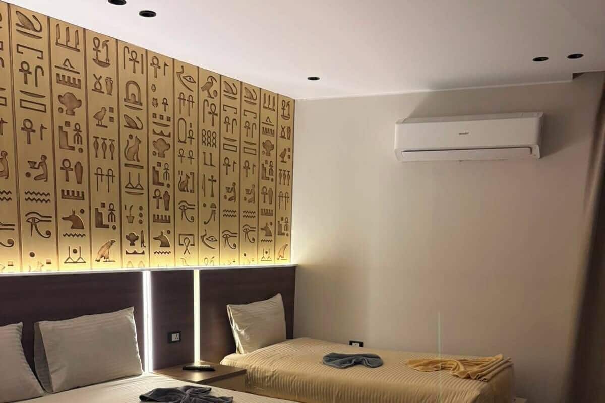 A hotel room with two beds, a wall with hieroglyphs, a ceiling light, and an air conditioning unit.