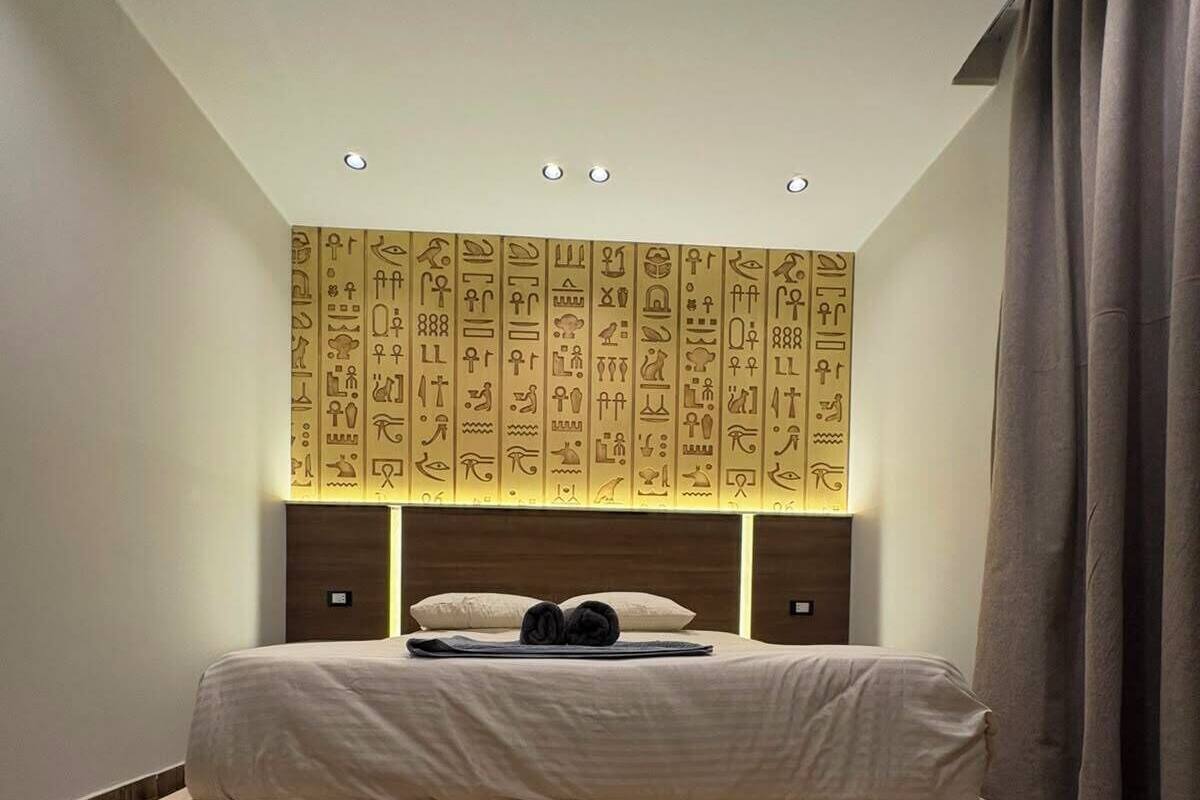 A bedroom with a bed, a wall with hieroglyphs, a rug, and a pair of shoes.