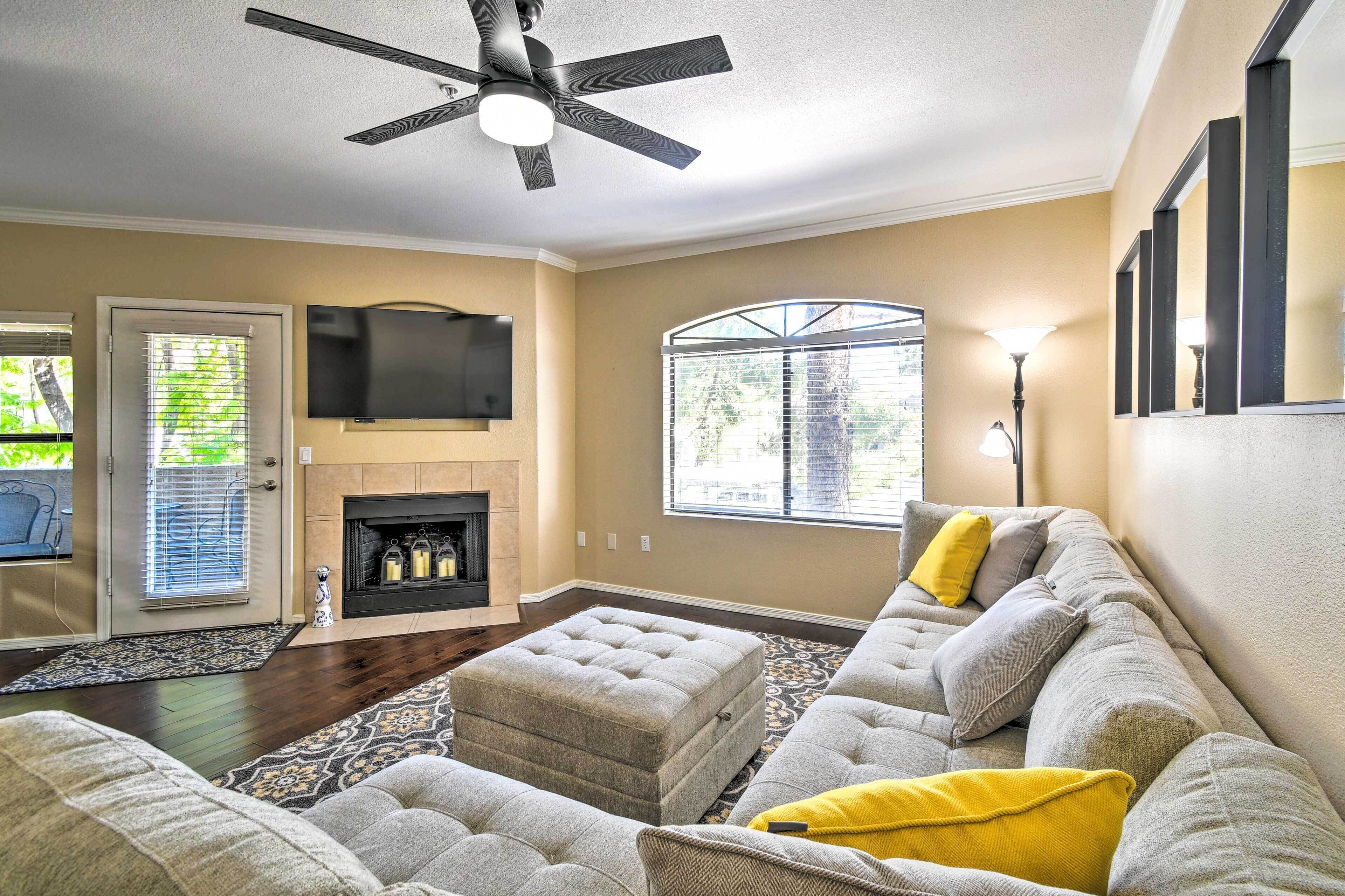 A living room with a fireplace, a ceiling fan, a TV mounted on the wall, and a large window with blinds.