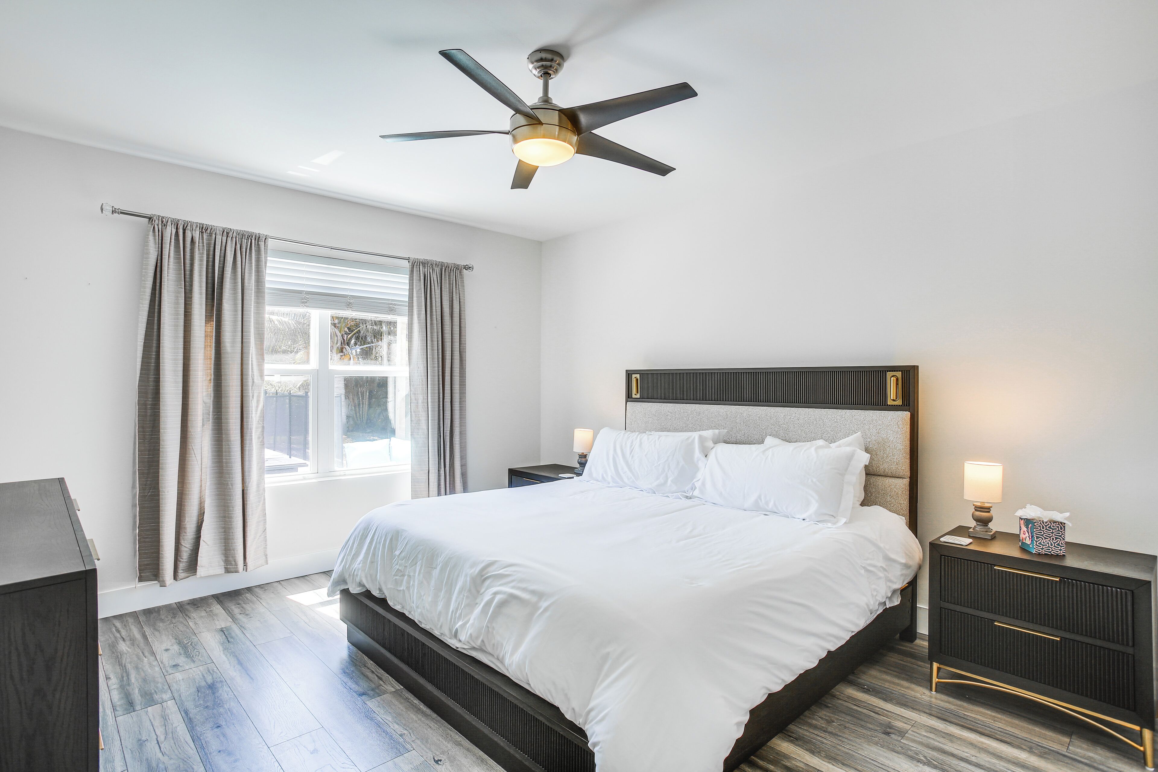 A bedroom with a bed, a ceiling fan, a nightstand with a lamp, and a window with curtains.