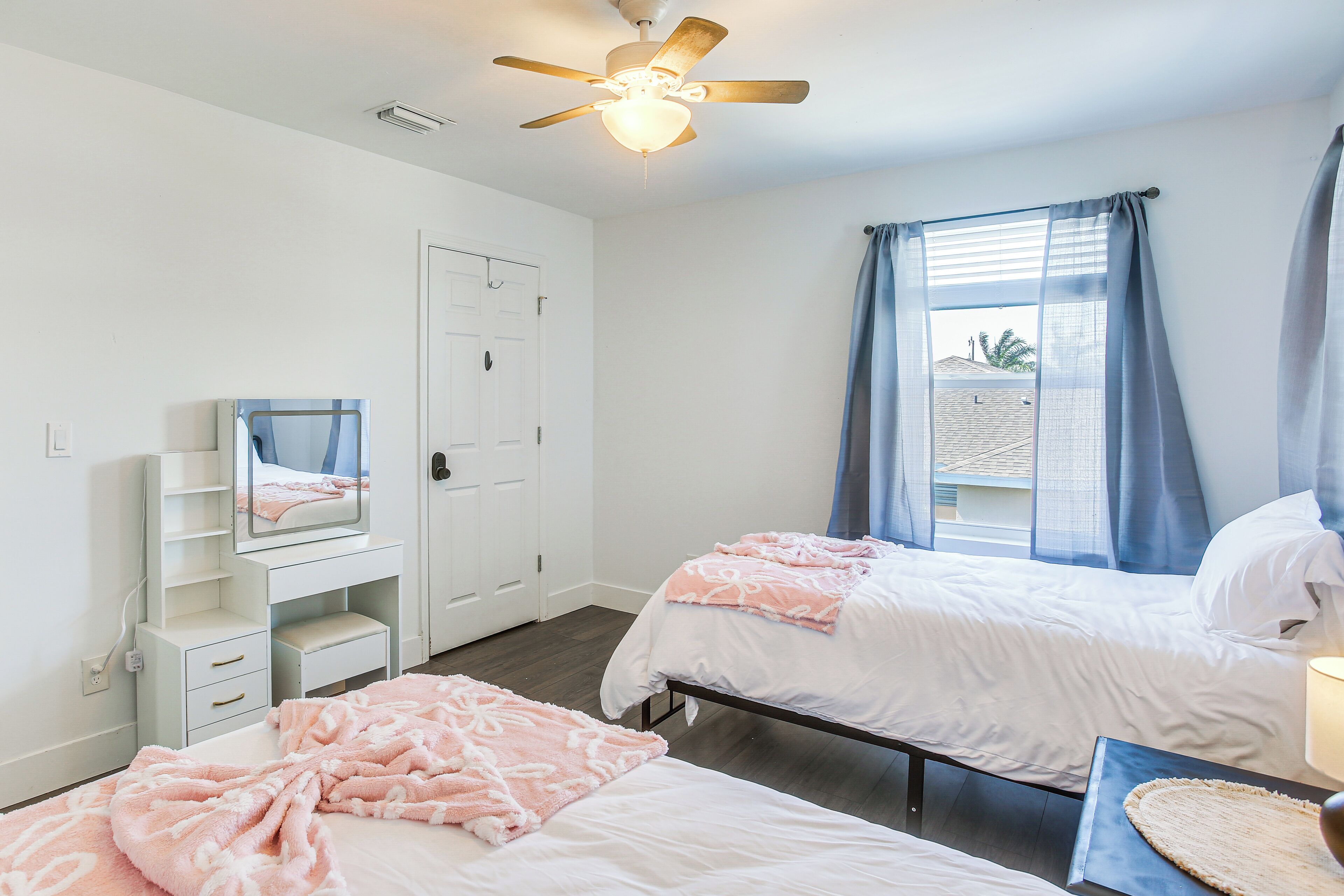 A bedroom with two beds, a ceiling fan, a window with curtains, a desk, and a mirror.