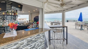 A bar area with a tiled backsplash, a flat-screen TV, and a variety of bottles. There are outdoor tables and chairs, and a blue umbrella.