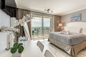 Quarto de hotel com cama, escrivaninha, cadeira, planta em vaso, janela com cortinas e um quadro na parede.