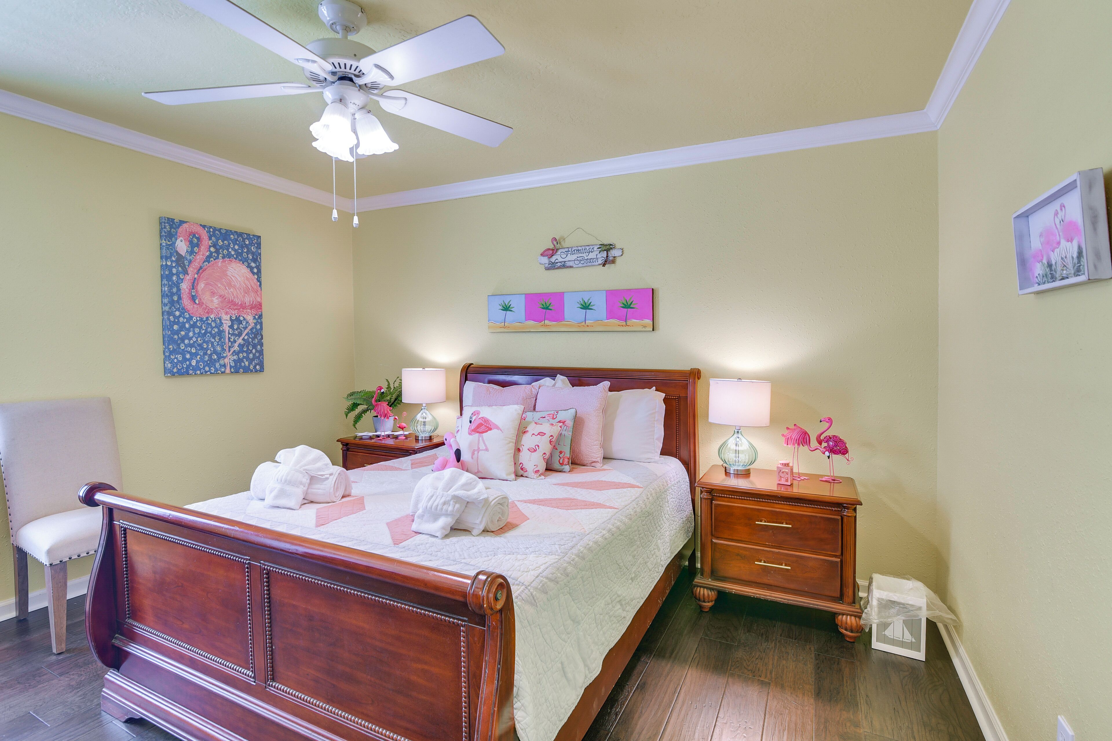 A bedroom with a wooden bed, a ceiling fan, a nightstand with a lamp, and a wall with a painting and a sign.