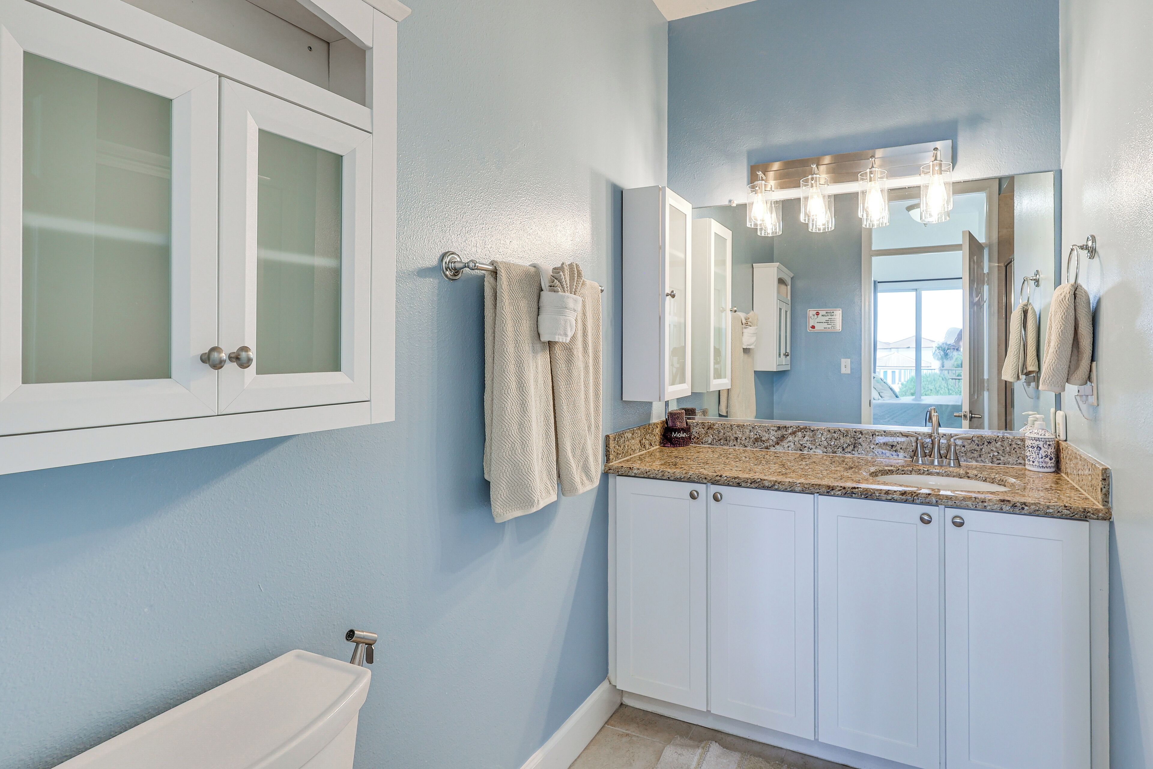 A bathroom with a white toilet, a sink with a granite countertop, a mirror with lights, and a towel rack.