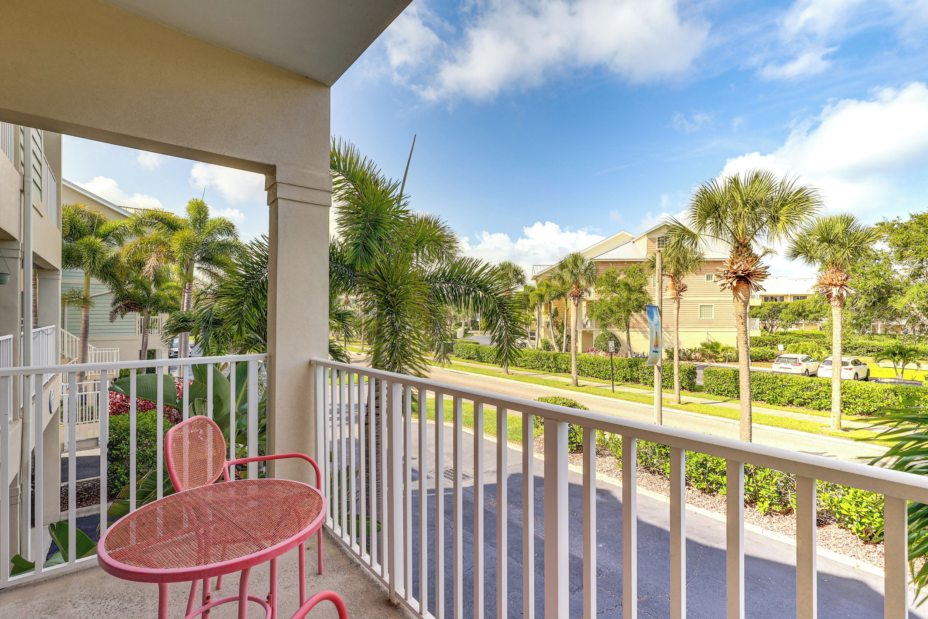 A balcony with a white railing, two pink chairs, and a view of a street with palm trees and houses.