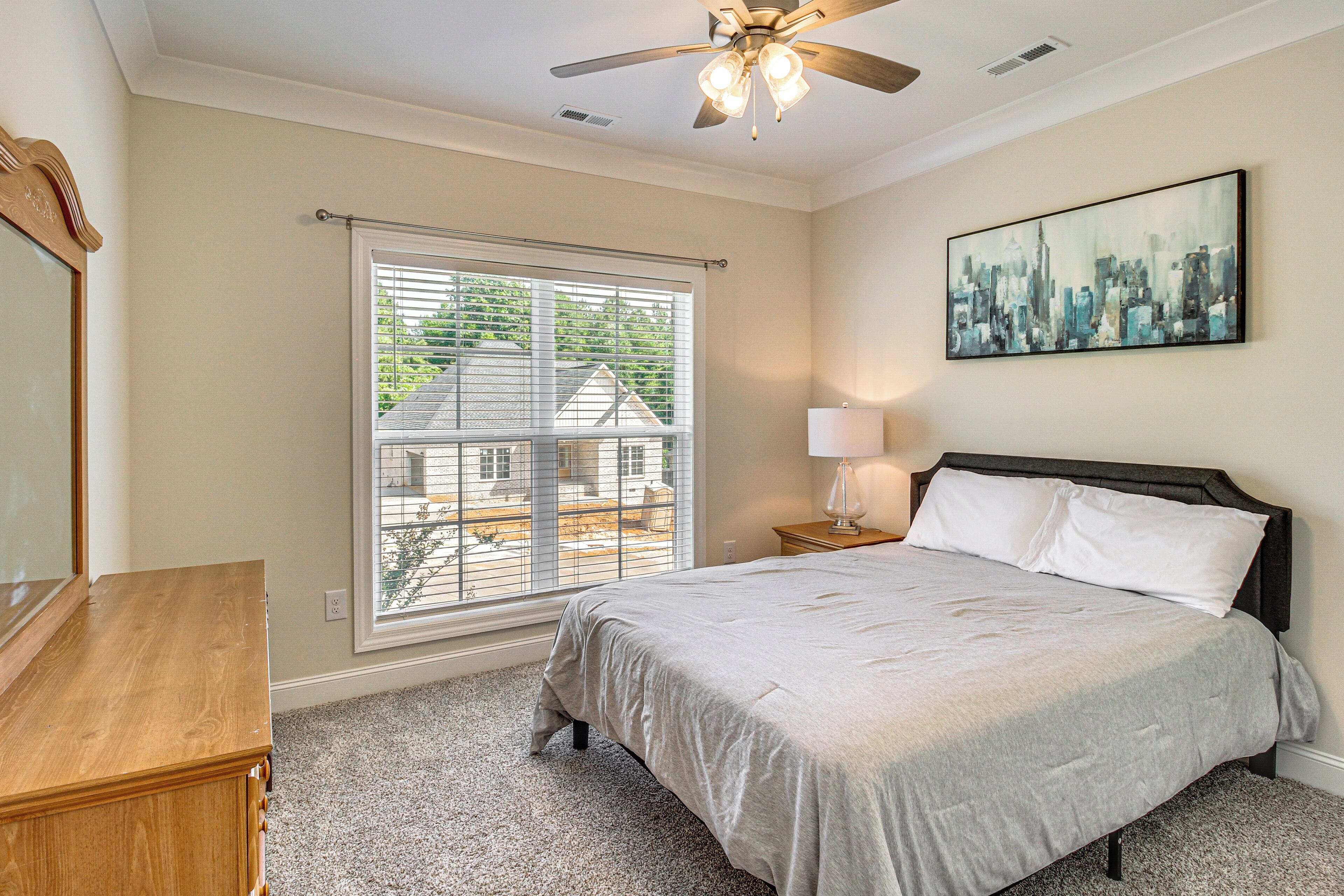 A bedroom with a bed, a nightstand, a lamp, a ceiling fan, a window with blinds, and a cityscape painting on the wall.