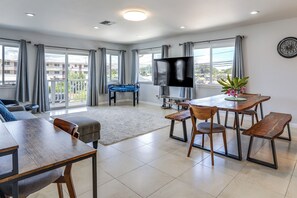 A spacious living area with a sofa, a dining table with benches, a foosball table, a flat-screen TV, and large windows with curtains. - Walk to Sand! Ewa Beach Group Retreat (Ewa Beach)