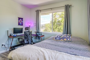 Un dormitorio con una cama, un escritorio con computadora, una silla y un ventanal con cortinas.