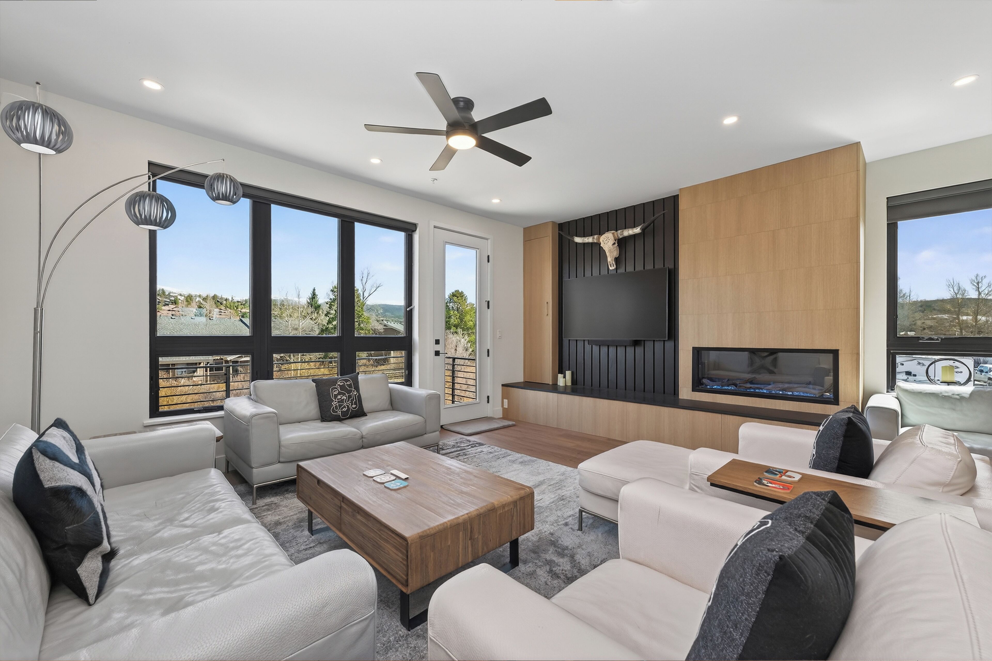 A modern living room with a sofa, coffee table, and TV, large windows, and a ceiling fan.