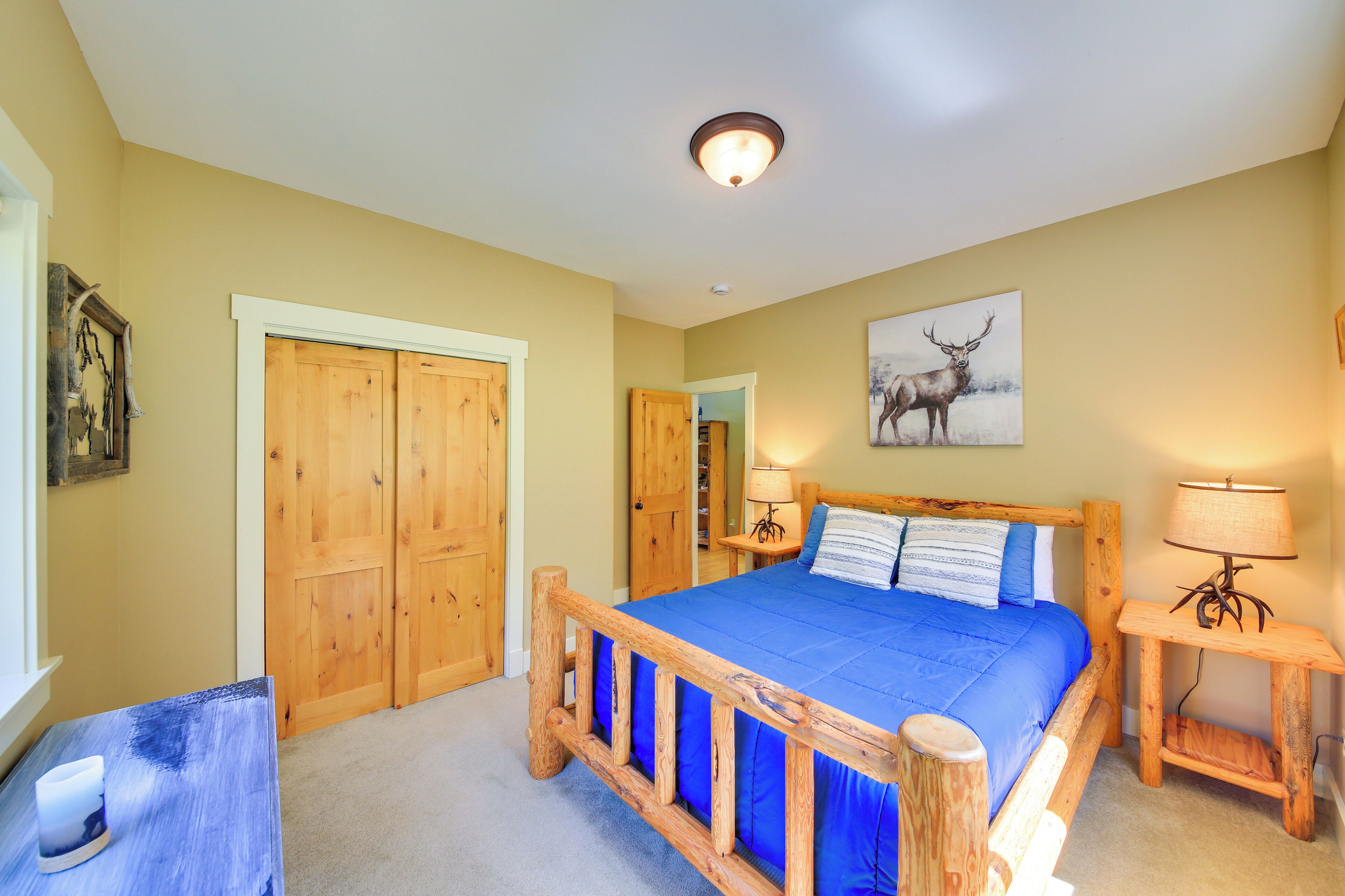 A bedroom with a wooden bed frame, a blue bedspread, two bedside lamps, and a deer painting on the wall.