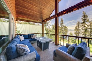 A covered outdoor seating area with a pool table, blue sofas, and a view of trees and water. - Lake Access + Water Views: Bottle Bay Family Home! (Sagle)