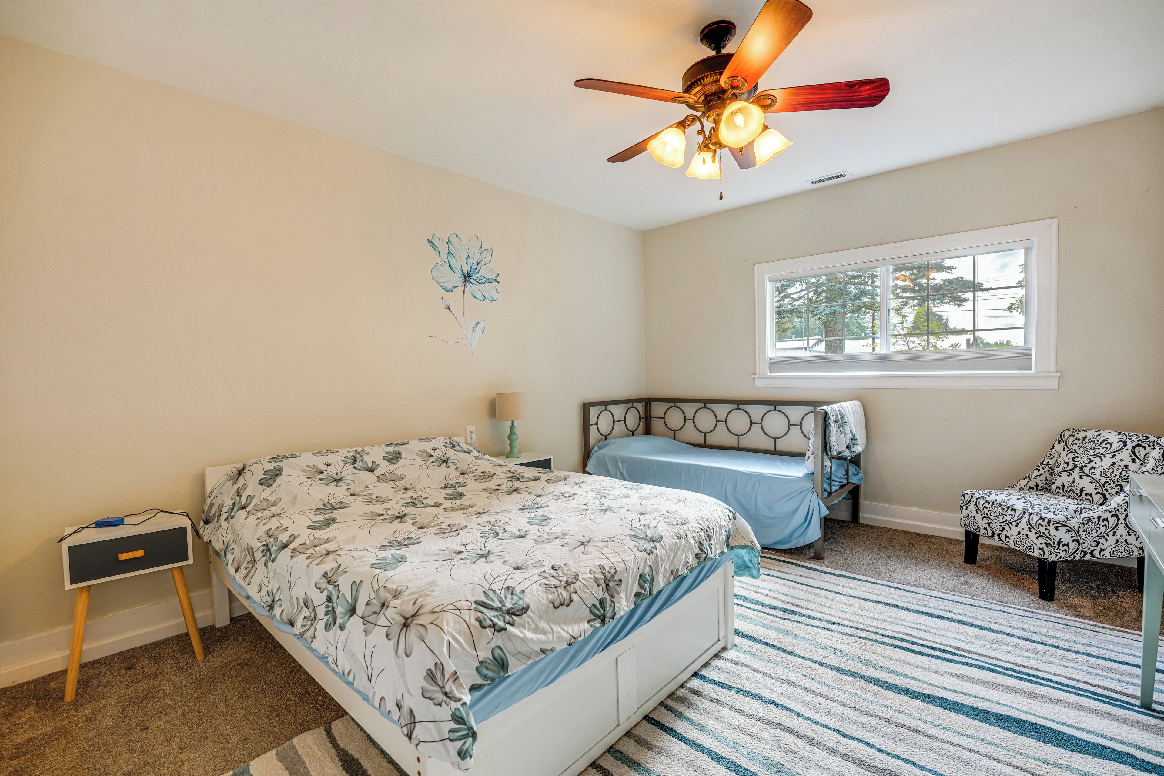 A bedroom with a bed, a desk, a chair, a ceiling fan, a window, and a wall decoration.