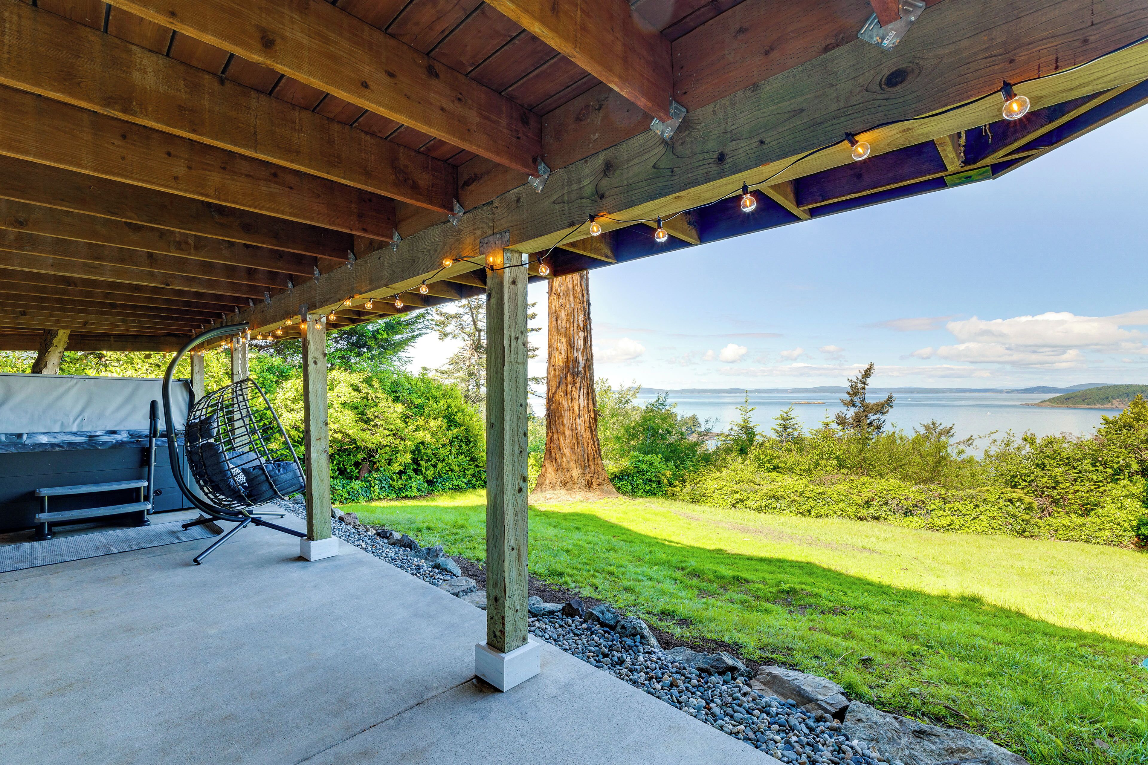 Un espacio al aire libre techado con piso de concreto, una silla colgante y vista a un lago y bosque.