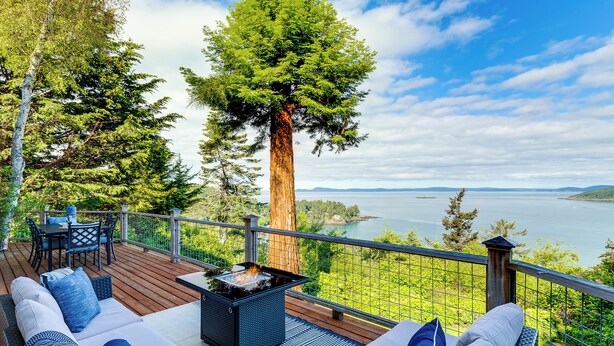 A deck with outdoor seating, a fire pit, and a view of a lake and forest. - Snow Goose Point: Stunning Views & New Hot Tub! (Anacortes)