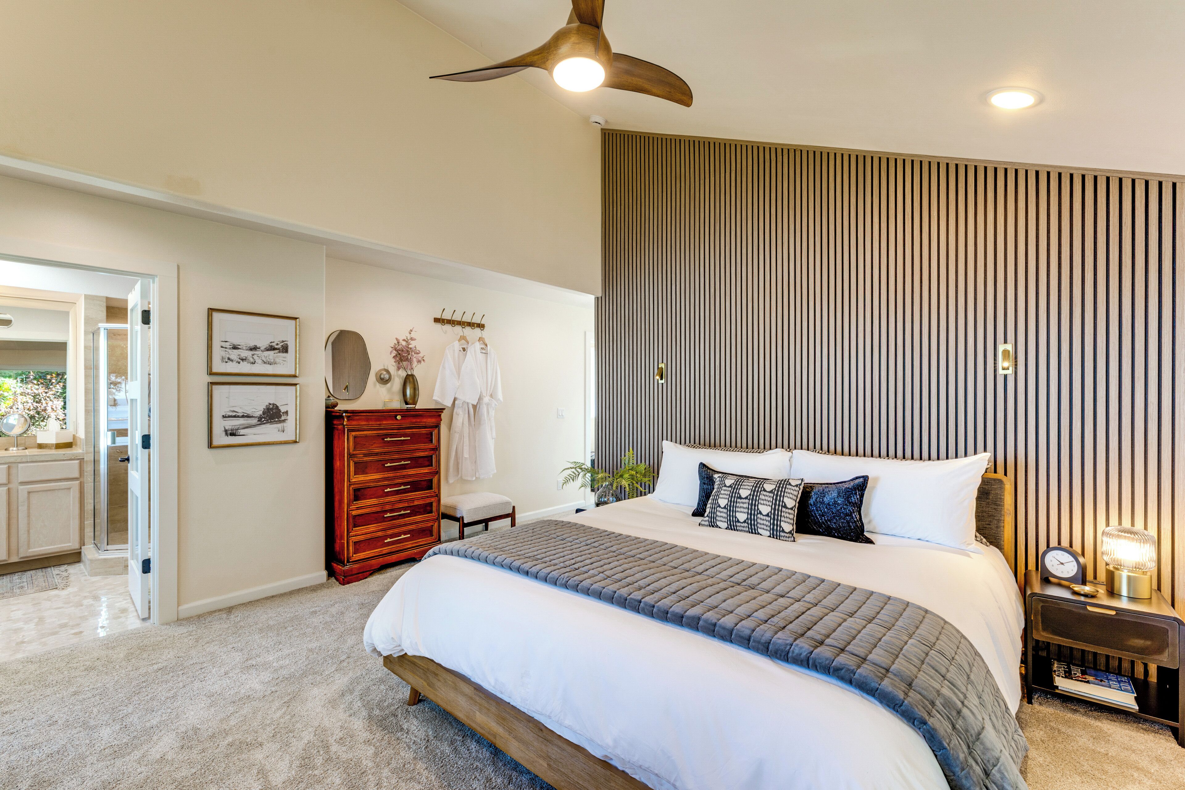 Een slaapkamer met een groot bed, een plafondventilator, een nachtkastje met lamp en een houten dressoir.