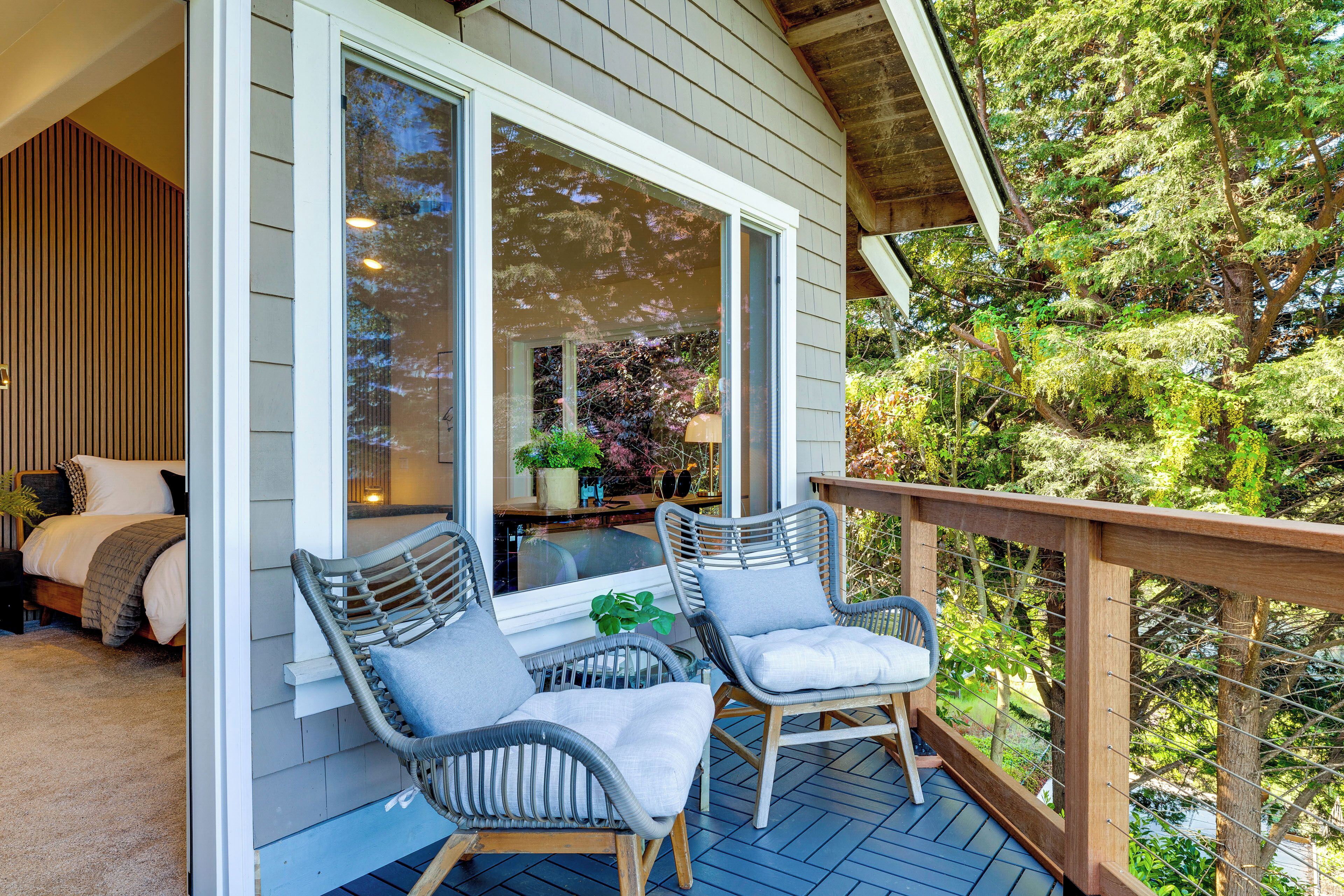 Une terrasse en bois avec des chaises en osier, une porte vitrée donnant sur une pièce avec un lit et une plante, et une vue sur les arbres.