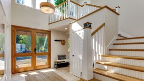 A staircase with wooden steps and white railings, a door, and a bench in a bright room with large windows. - Snow Goose Point: Stunning Views & New Hot Tub! (Anacortes)