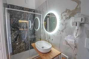 Un baño moderno con cabina de ducha de vidrio, un lavabo blanco sobre una cómoda de madera y un espejo grande con iluminación integrada.
