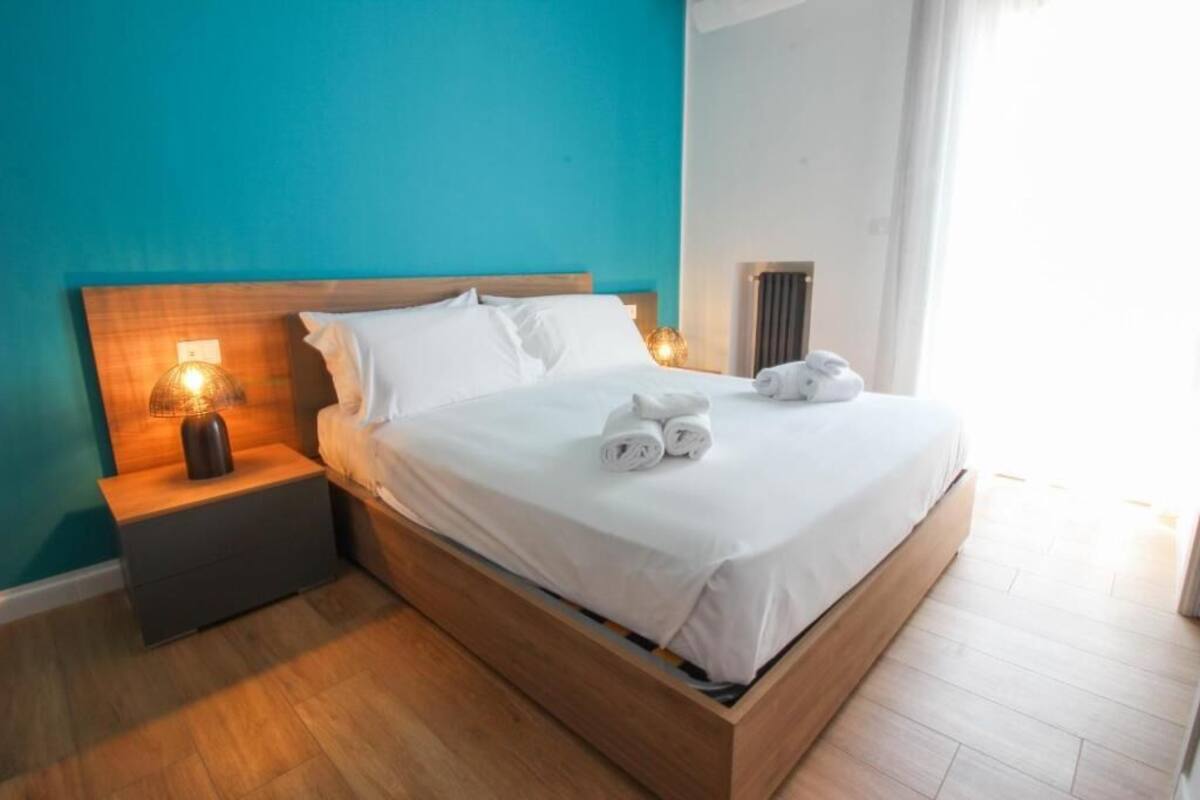 Una cama bien hecha con sábanas blancas y toallas enrolladas, una lámpara de noche y un cabecero de madera.