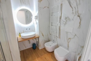 Un baño con pared de mármol, un espejo redondo con iluminación, un lavamanos, un bidé y un inodoro.