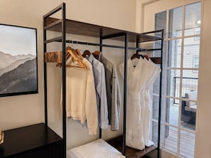 A clothing rack with various garments, a bed with a white pillow, and a window with a view of a building and balcony.