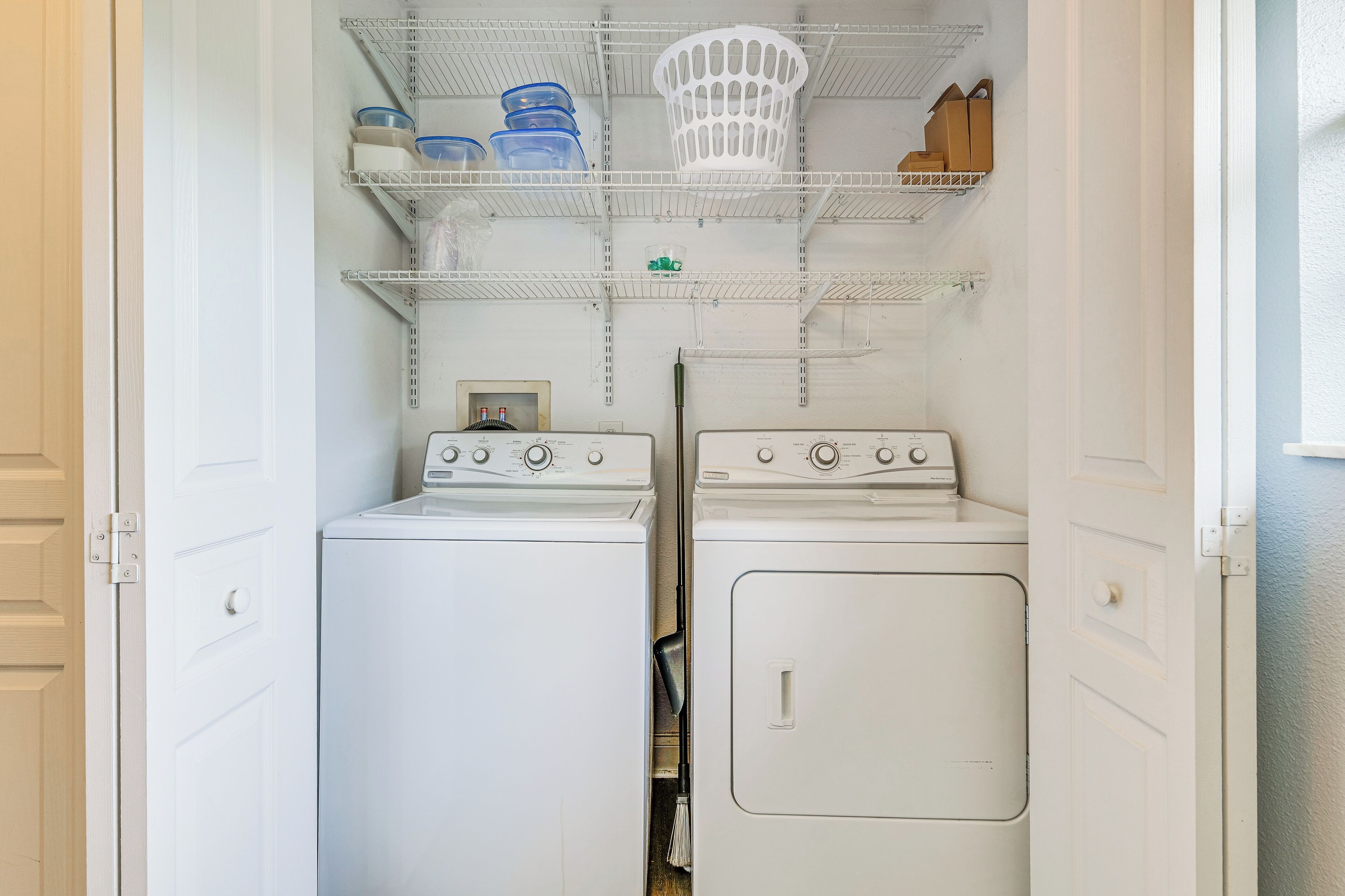 A laundry room with two washing machines, a dryer, and shelves above holding various containers and a basket.