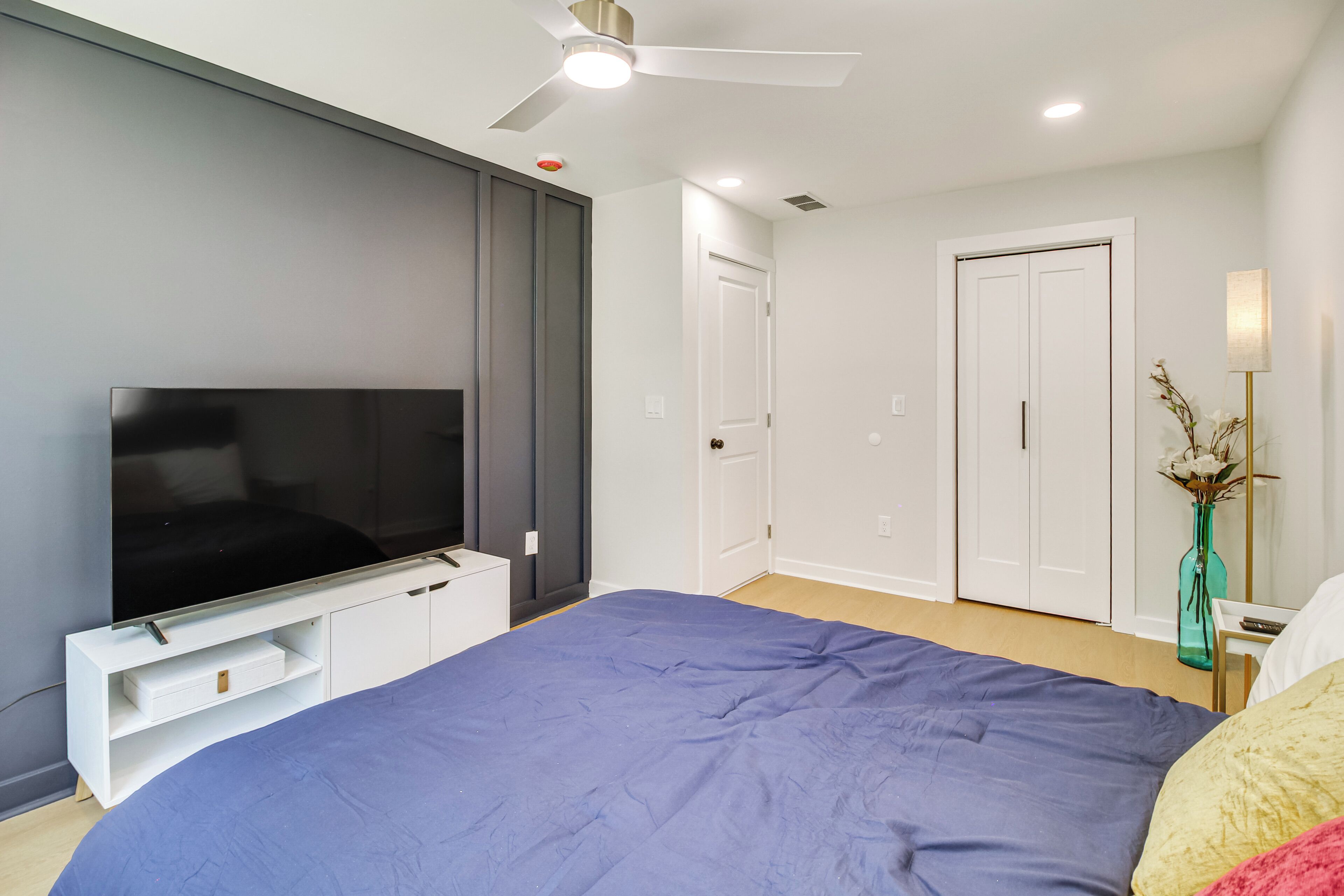 A bedroom with a bed, a TV, a ceiling fan, and a door.