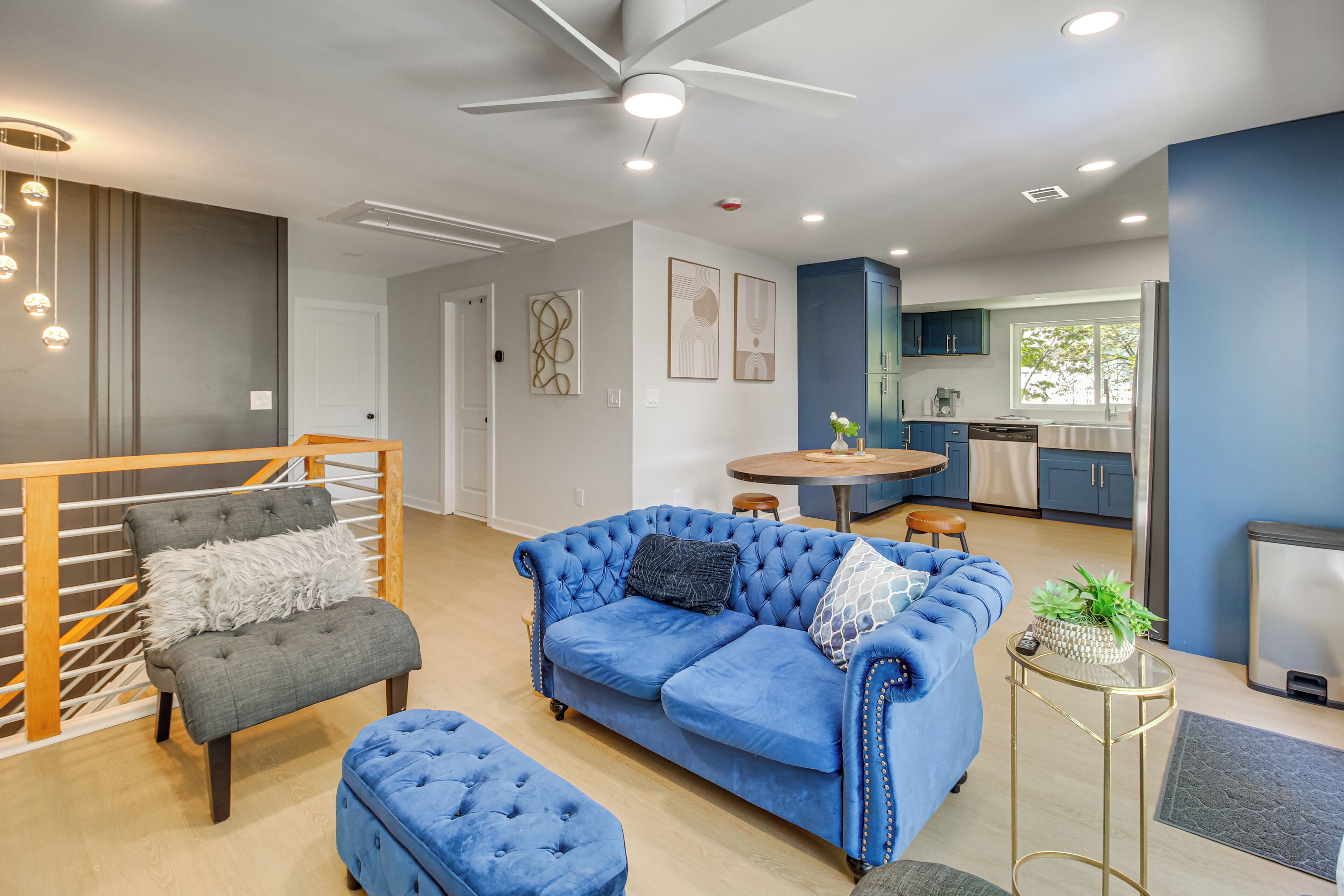 Una sala moderna con un sofá azul, un sillón gris y un reposapiés, un comedor con una mesa redonda y una cocina con armarios azules y un refrigerador.