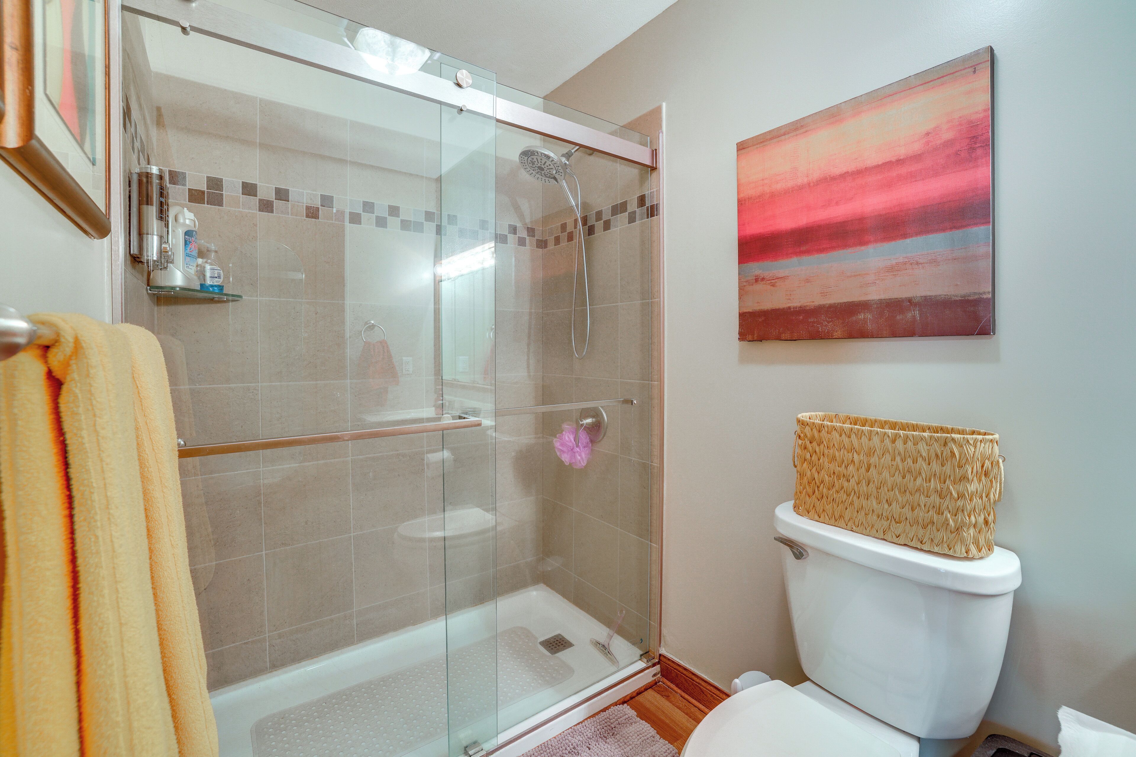 A bathroom with a glass-enclosed shower, a white toilet, a woven basket on the toilet tank, and a pink flower on the wall.