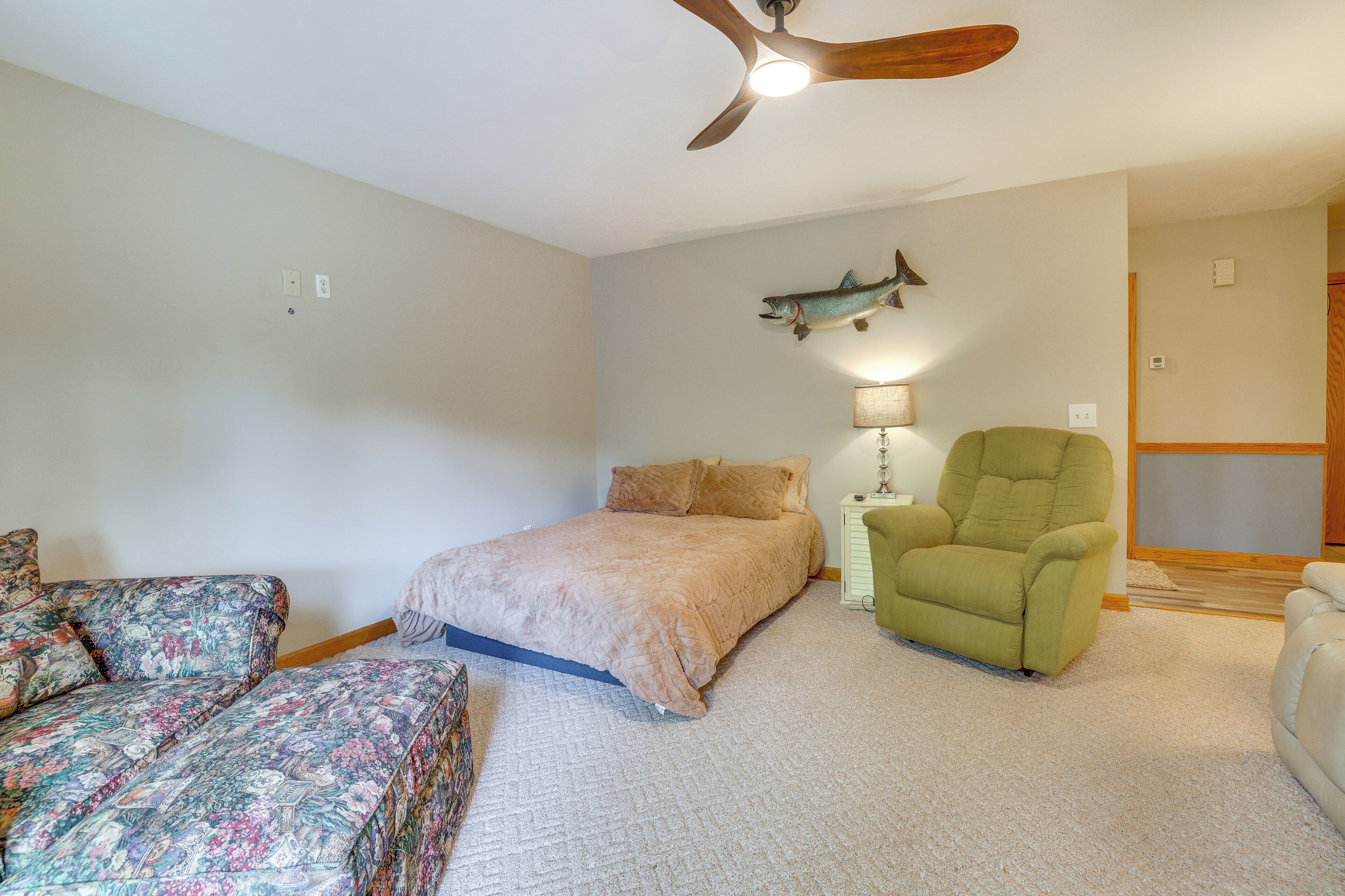 A bedroom with a bed, a green armchair, a patterned sofa, a ceiling fan, and a fish decoration on the wall.