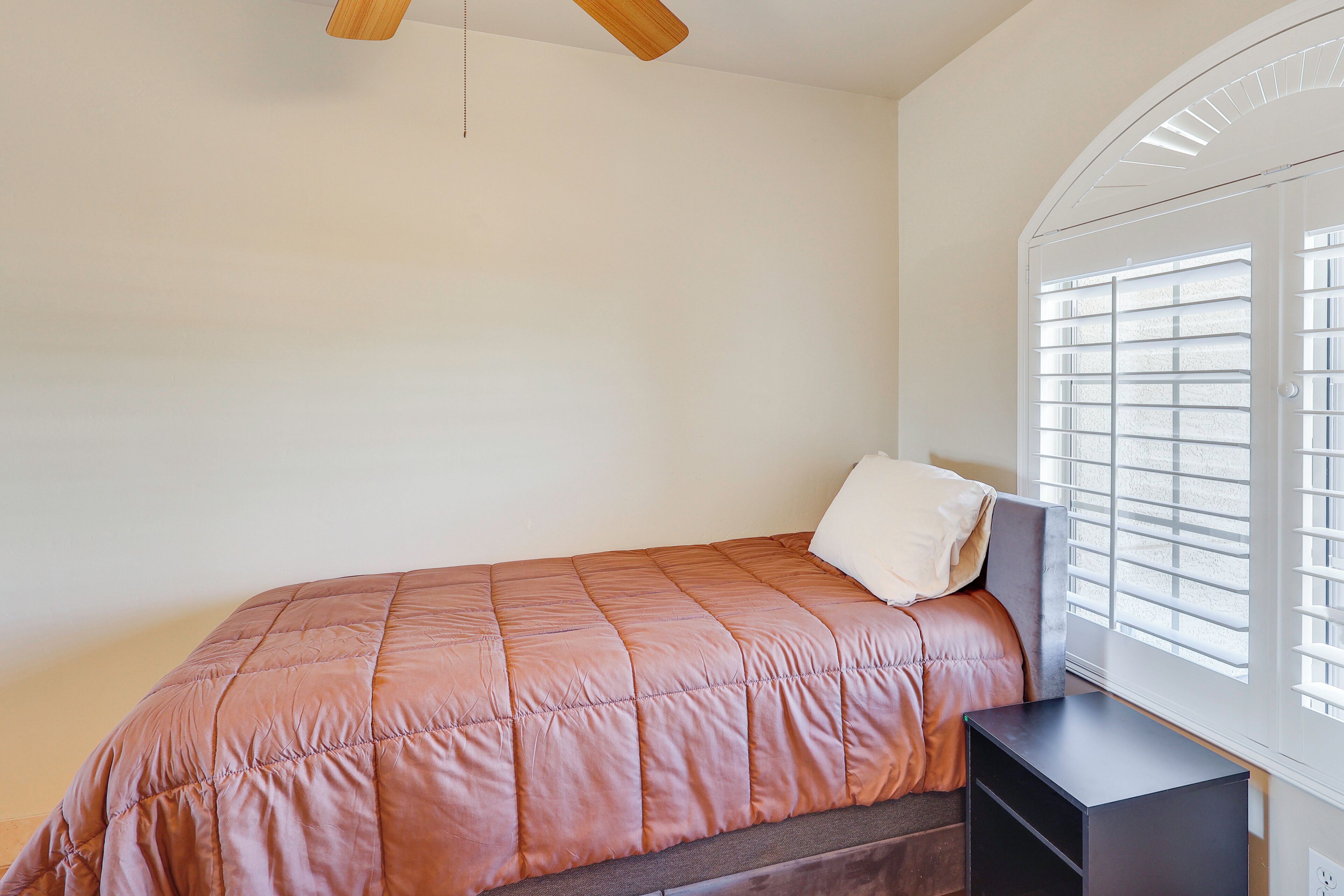 A bedroom with a bed, a bedside table, a window with blinds, and a ceiling fan.