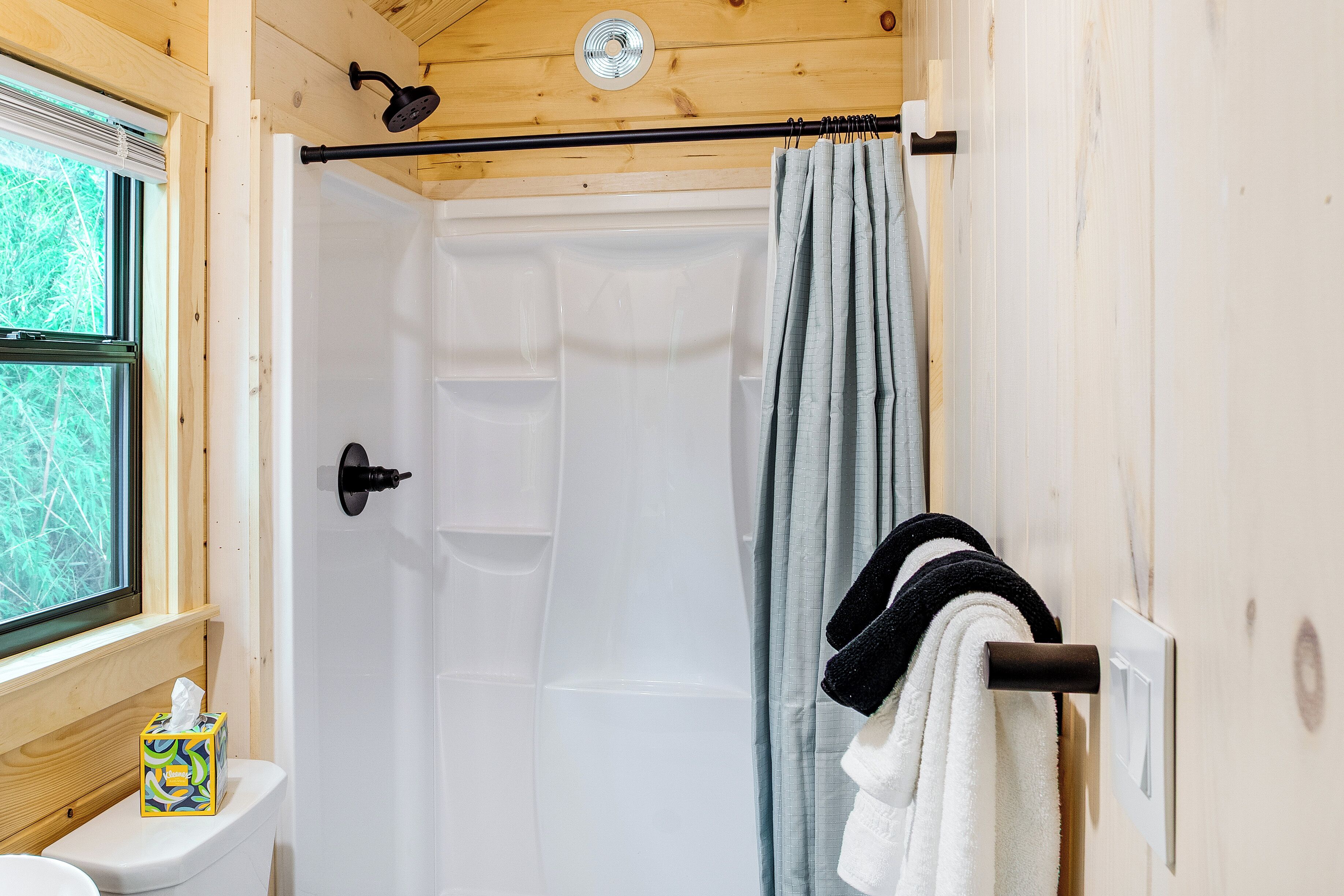 A shower with a curtain, a window, a towel rack with towels, and a toilet with a tissue box.