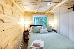 Un dormitorio de cabaña de madera con una cama, almohadas, una ventana y una mesita de noche.
