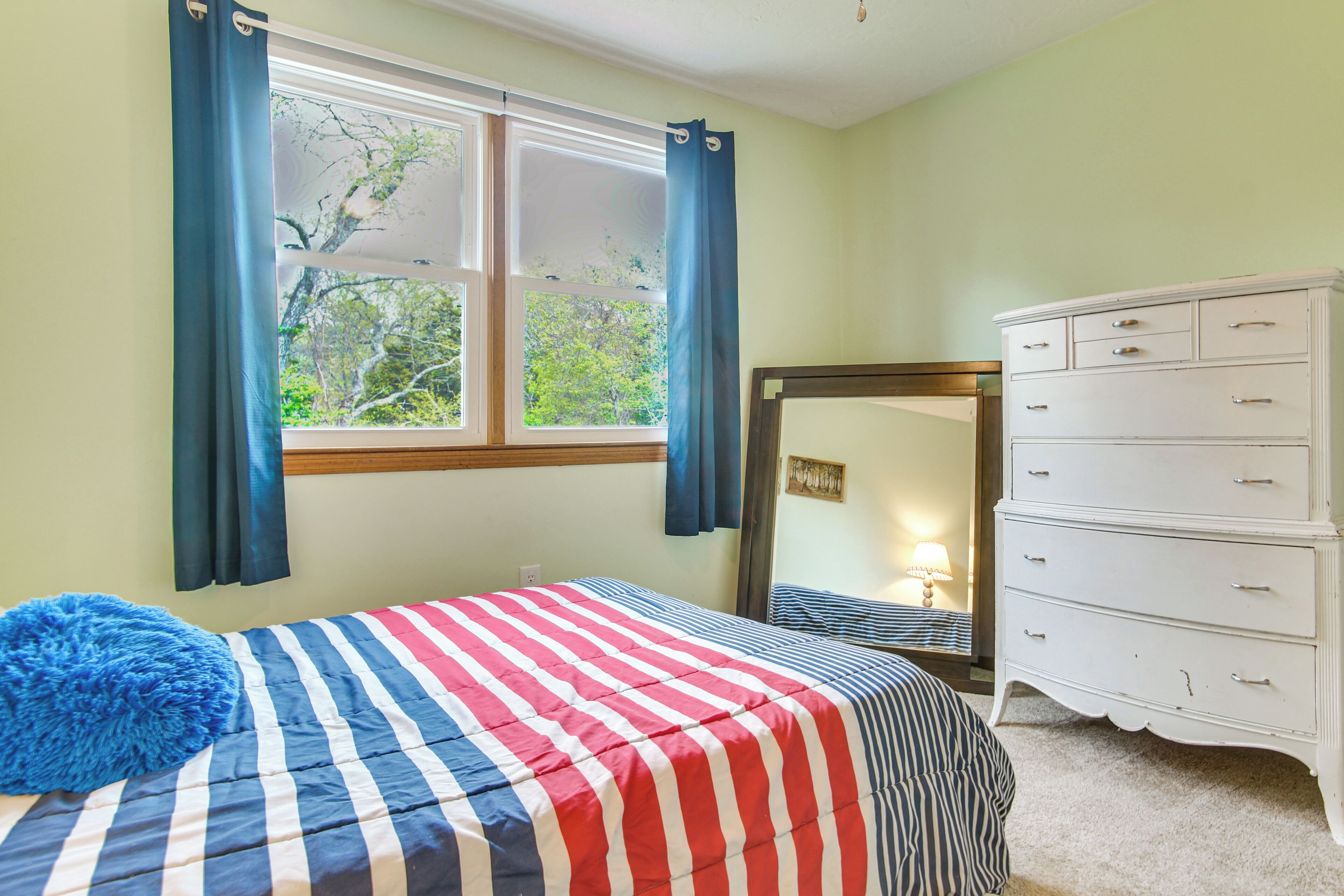 A bedroom with a bed, a dresser, a window with curtains, and a view of trees outside.