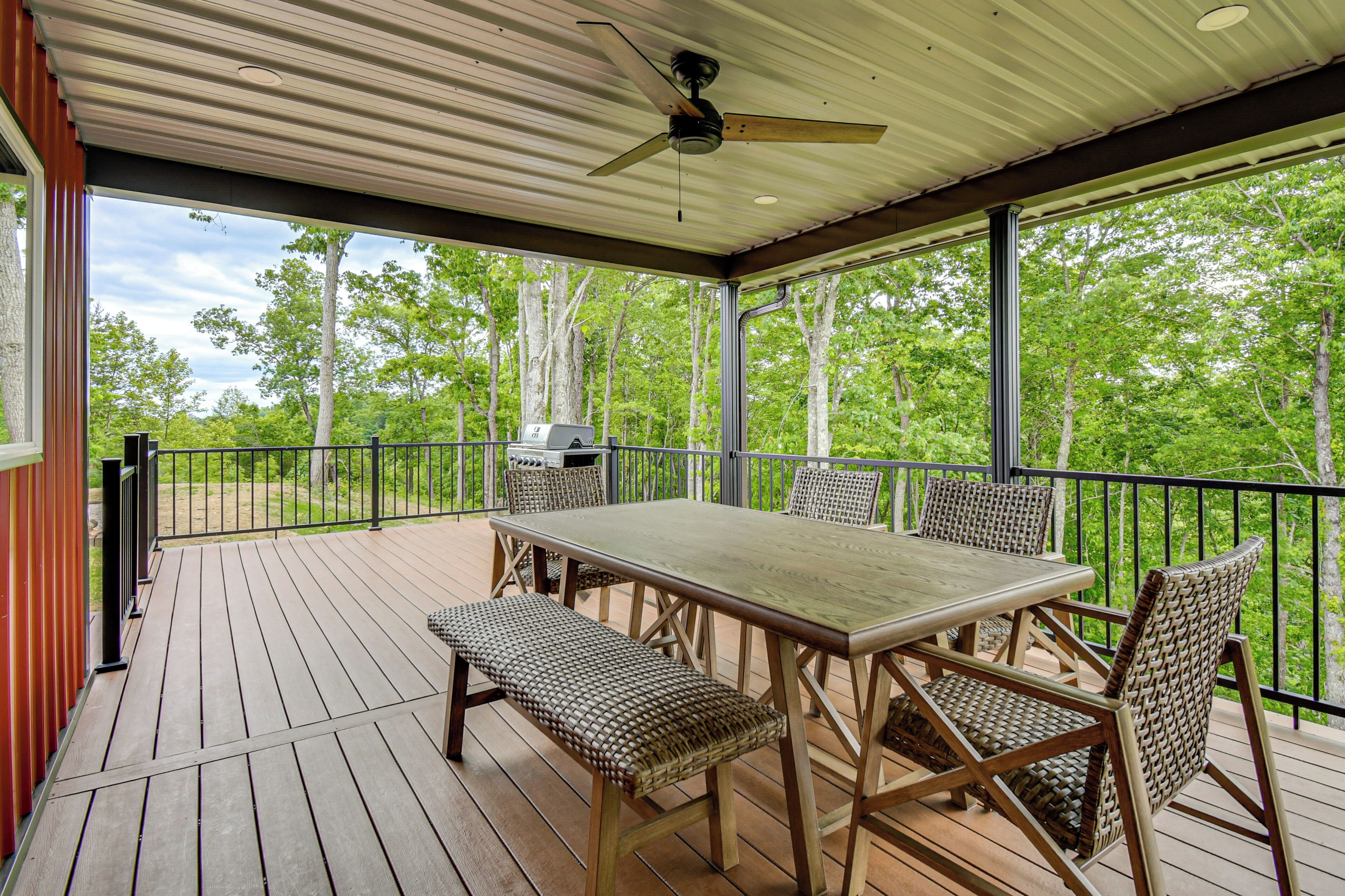 Une terrasse extérieure couverte, dotée d’une table et de chaises, d’un ventilateur de plafond et offrant une vue sur les arbres.