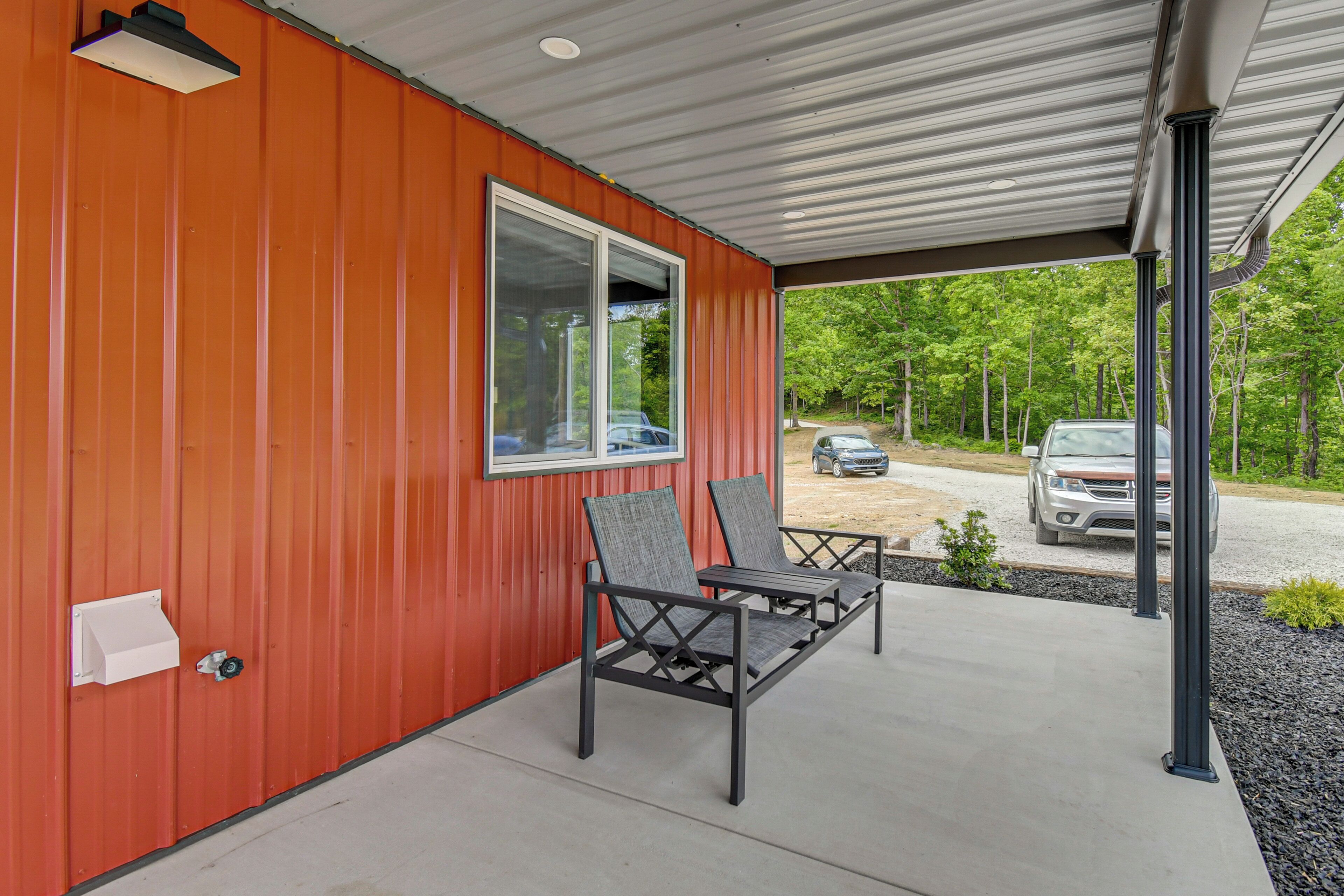 Un espace extérieur abrité avec un bâtiment rouge, deux chaises et une vue sur un parking.