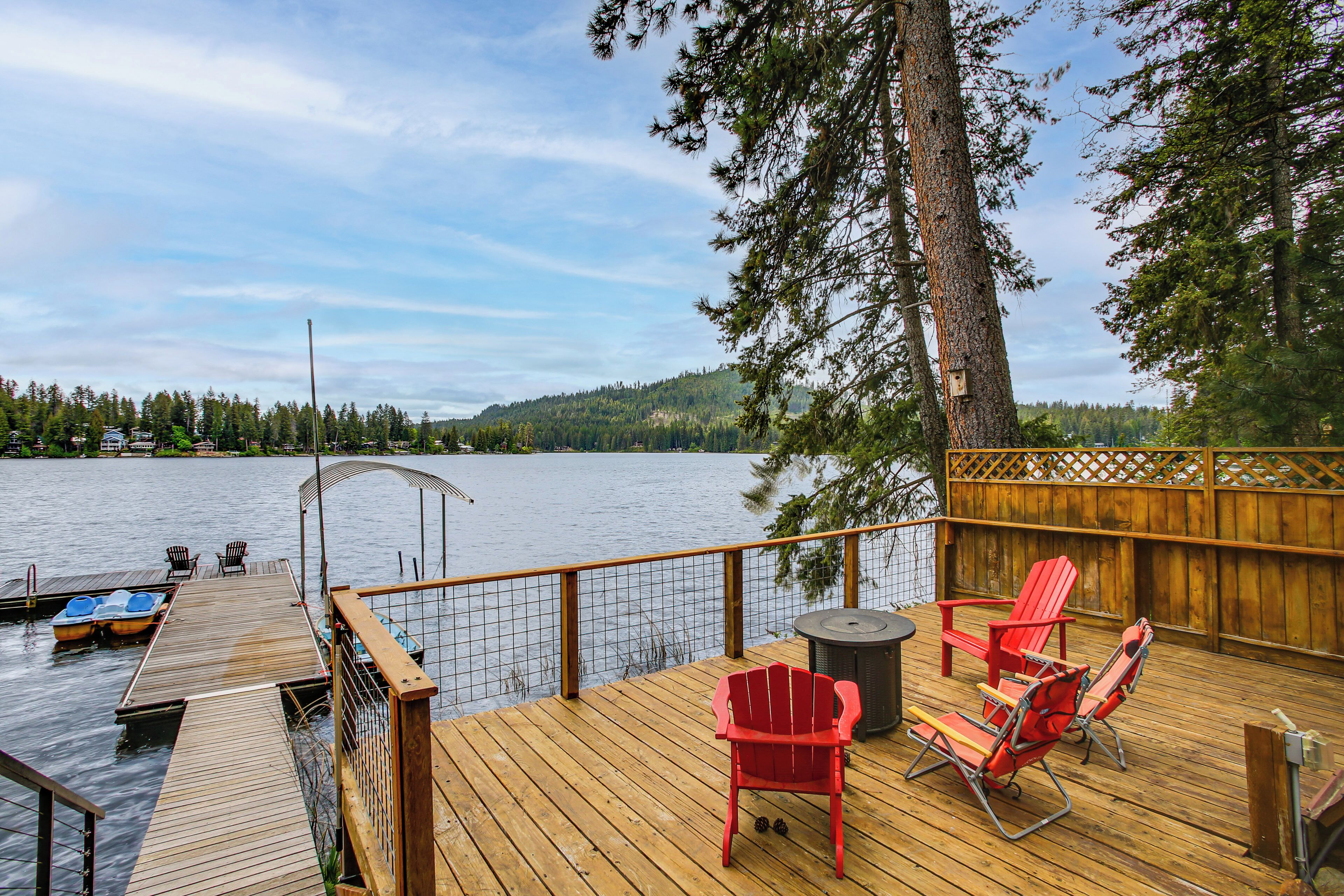 A wooden deck with a view of a lake, red Adirondack chairs, and a small table.
