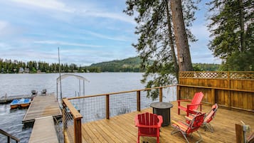 Una terraza de madera con vista a un lago, sillas Adirondack rojas y una mesita.