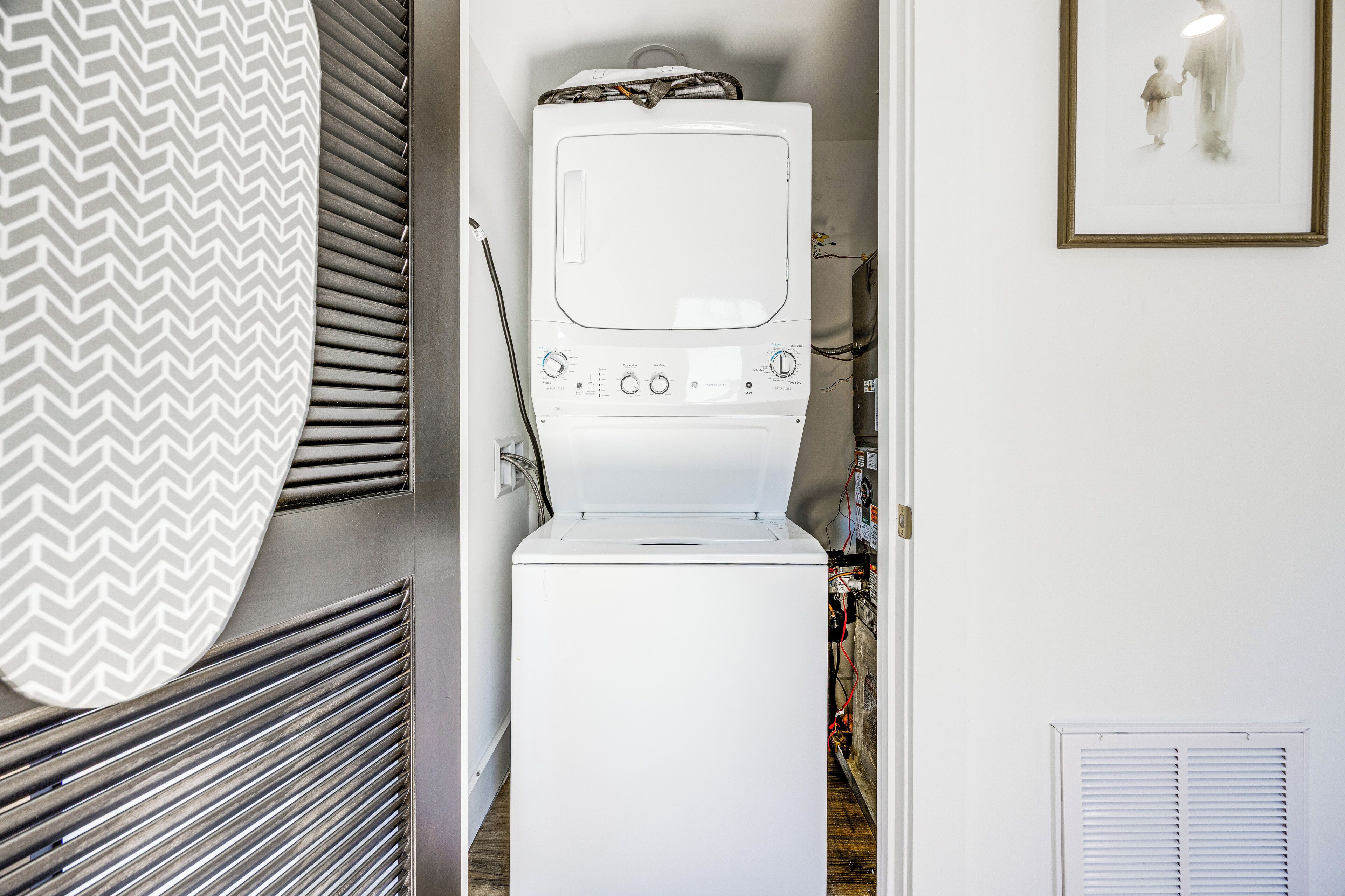 Uma lavanderia compacta com máquina de lavar e secadora empilhadas, um duto de ventilação na parede e um quadro.