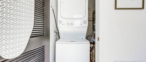 Uma lavanderia compacta com máquina de lavar e secadora empilhadas, um duto de ventilação na parede e um quadro.
