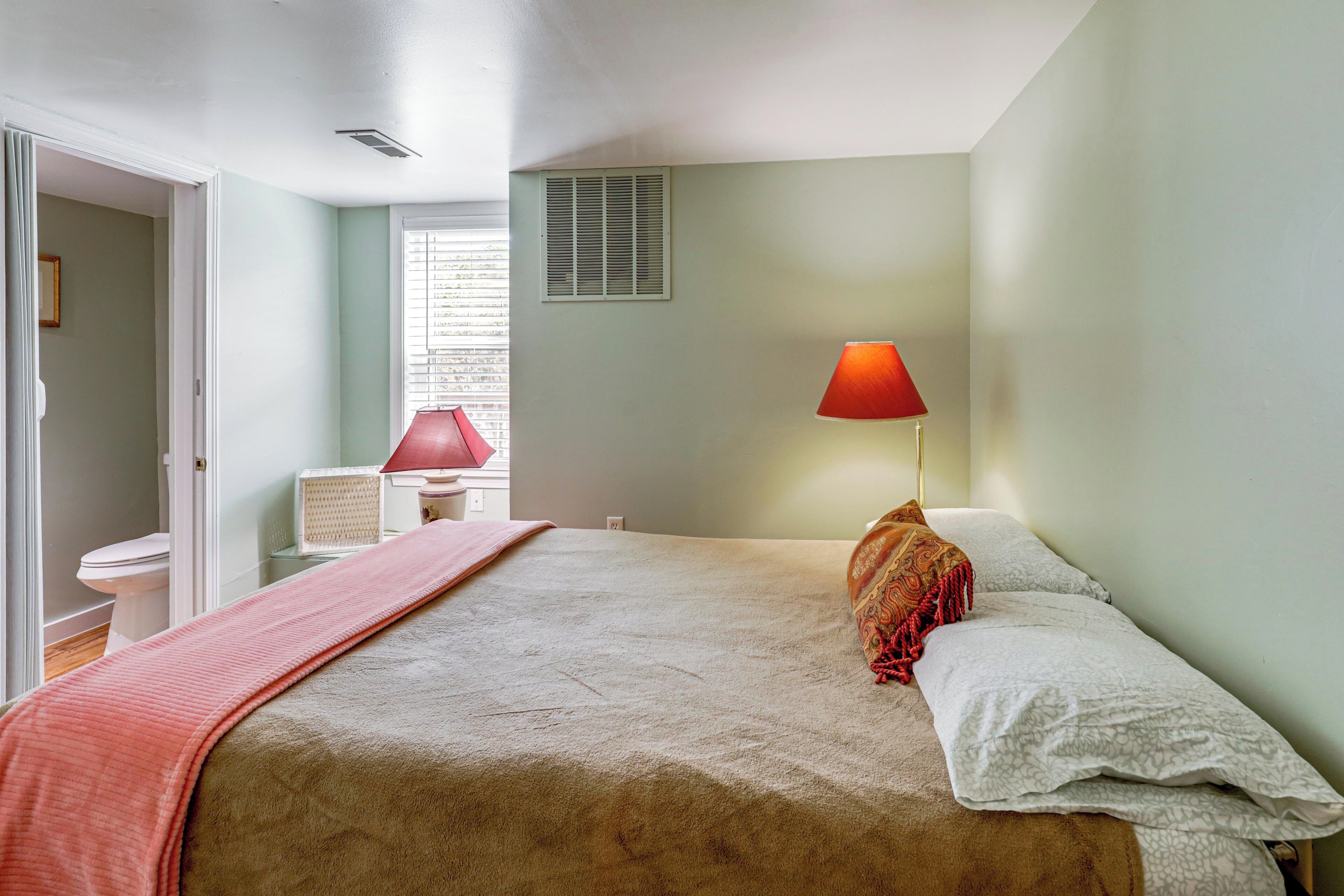 A bedroom with a bed, a bedside lamp, a window with blinds, and a door leading to another room.