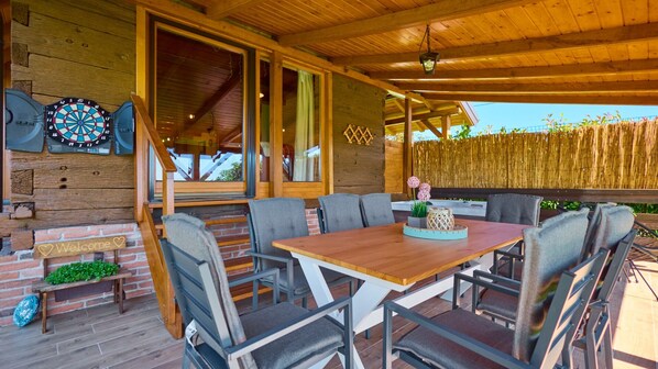 A covered outdoor seating area with a wooden table and chairs, a dartboard, and a welcome sign.