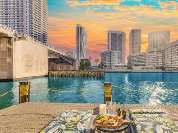 A waterfront view with high-rise buildings, a dock, and a dining setup with a plate of food and drinks. - Marina Front - 2BR - Beachwalk Resort (Hallandale Beach)
