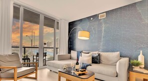 A modern living room with a large window, a sofa, a coffee table, and a map-themed wall. - Marina Front - 2BR - Beachwalk Resort (Hallandale Beach)