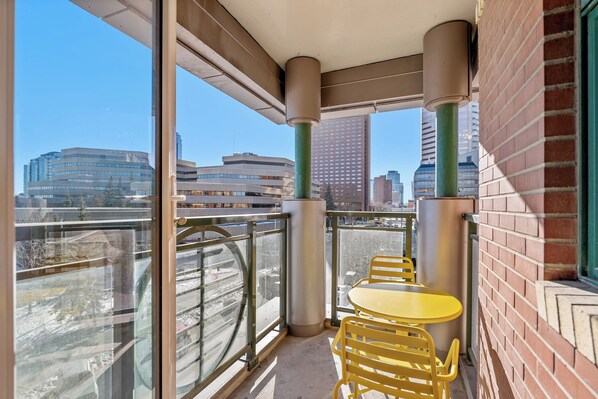 A balcony with a glass railing, a yellow table, and chairs, overlooking a cityscape with buildings. - 2 Bed, 2 Bath Condo in the Heart of Downtown Calgary Near Saddledome & Stampede (Calgary)